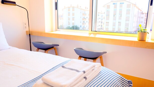 A hotel room with a bed, two chairs, a window with a view of buildings, and a plant on the windowsill. - ihW 1-bedroom Apartment Sunny & Comfy (Espinho)