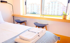 Ett hotellrum med en säng, två stolar, ett fönster med utsikt över byggnader och en växt på fönsterbrädan.