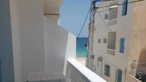 A balcony with a white table and chair, overlooking a clear blue sea and buildings with blue shutters.