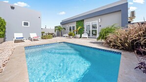 A modern pool area with a rectangular pool, white lounge chairs, and a grey building with a glass door. - 5 min Eagle Beach | New Luxury 1BR w/ Pool & BBQ (Noord)