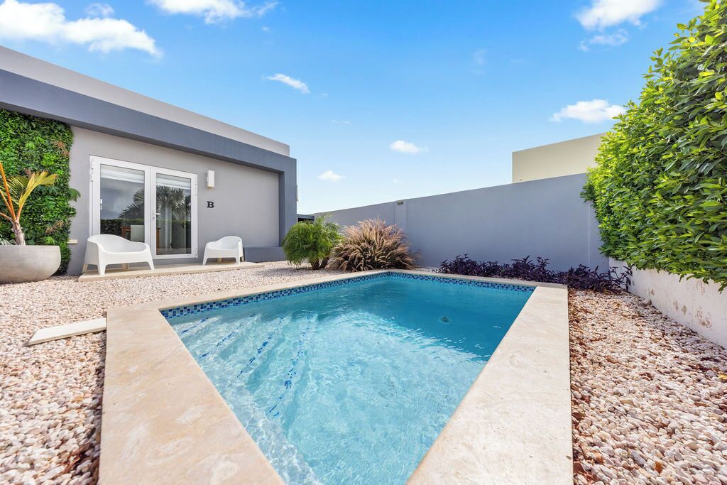 Una piscina con borde de azulejos, rodeada de piedras. Hay dos sillas blancas y una pequeña mesa cerca de un ventanal.