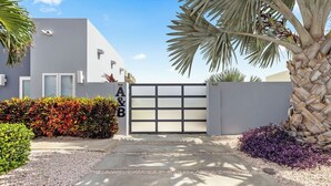 A modern building with a garage door, surrounded by gravel, a palm tree, and colorful foliage. - 5 min Eagle Beach | New Luxury 1BR w/ Pool & BBQ (Noord)
