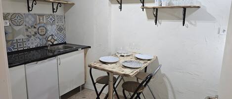 A compact kitchen with white cabinets, a black countertop, and a dining table set with plates and glasses. There are two wall-mounted shelves above the counter, one holding plates and the other holding cups and a potted plant.