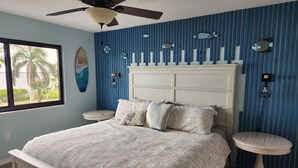 A bedroom with a bed, two bedside tables, a ceiling fan, and a window with a view of palm trees. - Harborfront Views, Rooftop Pool, walking distance to Fishermen's Village! (Punta Gorda)