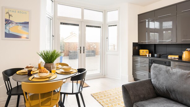 A modern kitchen with dark cabinets and a dining area with yellow chairs and a table set for breakfast. - Host Stay Ammonite (Lyme Regis)