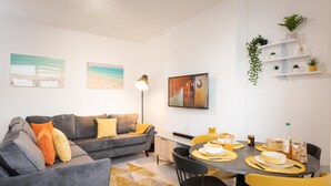 A modern living room with a grey sofa, yellow cushions, a dining table set with yellow placemats, and a mounted TV. - Host Stay Ammonite (Lyme Regis)