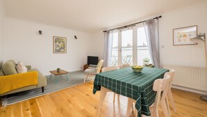 Ein Wohnzimmer mit einer Couch, einem Esstisch mit grüner Tischdecke, einem Fernseher und einem Fenster mit Vorhängen.