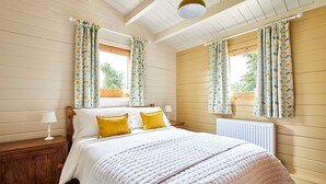 Un dormitorio con cama, cabecera de madera, dos mesitas de noche con lámparas y una ventana con cortinas.