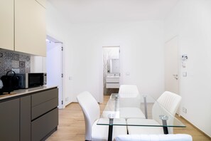 Una cocina moderna con microondas y cafetera, un comedor con mesa de vidrio y sillas blancas, y un baño con lavamanos y espejo blancos.