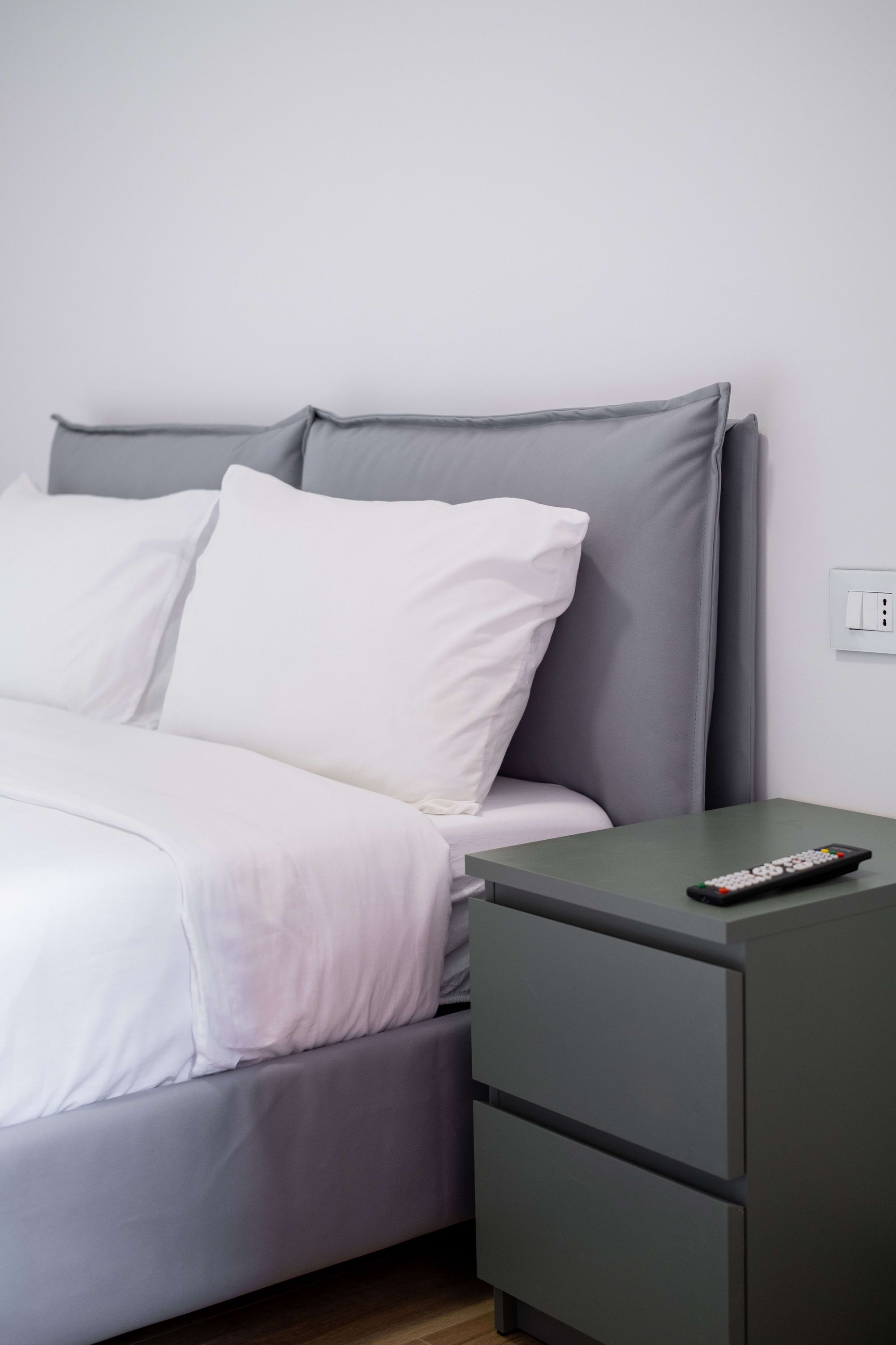 Un lit bien fait, avec une literie blanche et grise, une table de chevet verte avec une télécommande, et un mur blanc avec un interrupteur.