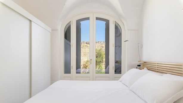 Ein Schlafzimmer mit Bett, weißen Wänden, einem Holz-Kopfteil und Blick durch eine Glastür in einen Außenbereich.
