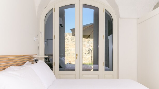 A bedroom with a bed, a wooden headboard, a white wardrobe with glass doors, and a view of a rocky outdoor area.