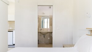 A bedroom with a bed, a nightstand, a bedside table, a wall-mounted shelf, and a bathroom visible through an open door. - Masseria Pichilicchio 1 by Wonderful Italy (Ostuni)
