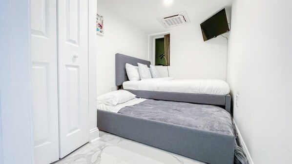 A compact bedroom with two beds, a white door, a mounted TV, and a small window.