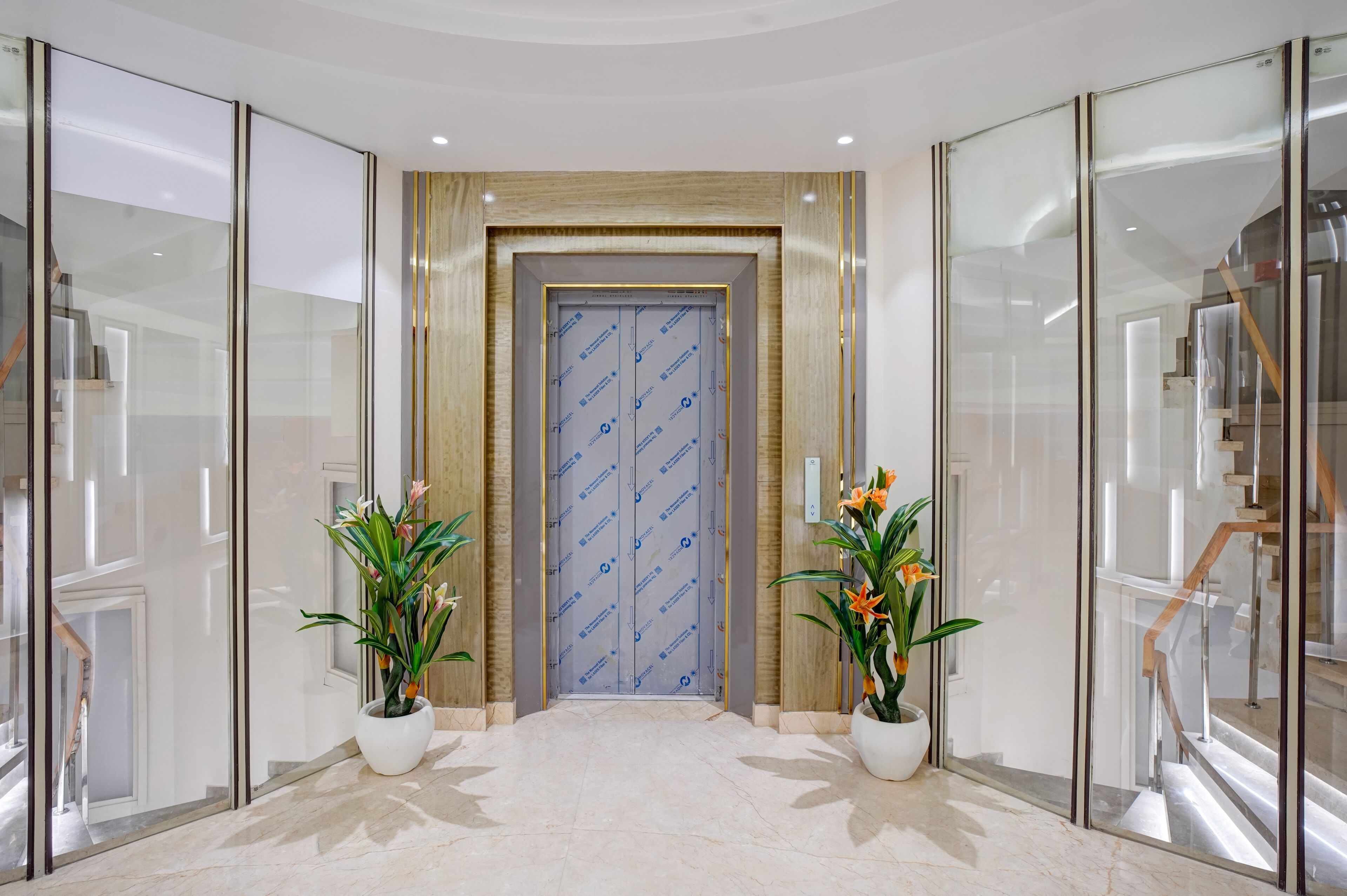 A modern hotel lobby with a glass elevator, marble flooring, and two potted plants.