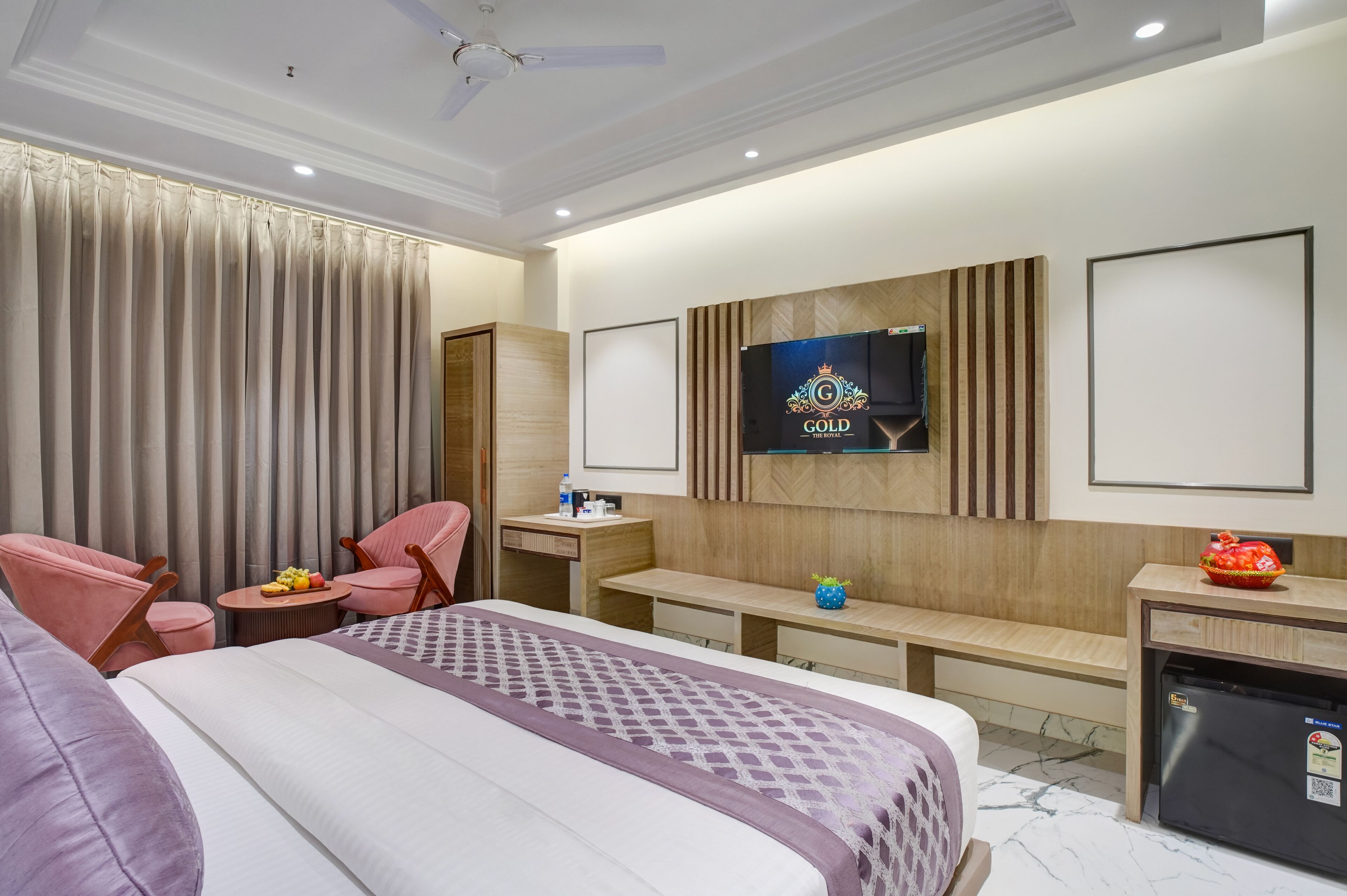 A hotel room with a bed, two pink chairs, a small table with fruit, a TV on the wall, a minibar, and a ceiling fan.
