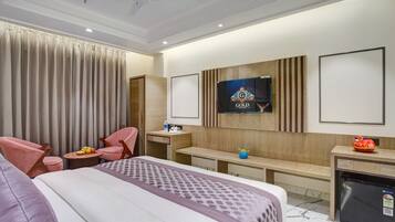 A hotel room with a bed, two pink chairs, a small table with fruit, a TV on the wall, a minibar, and a ceiling fan.