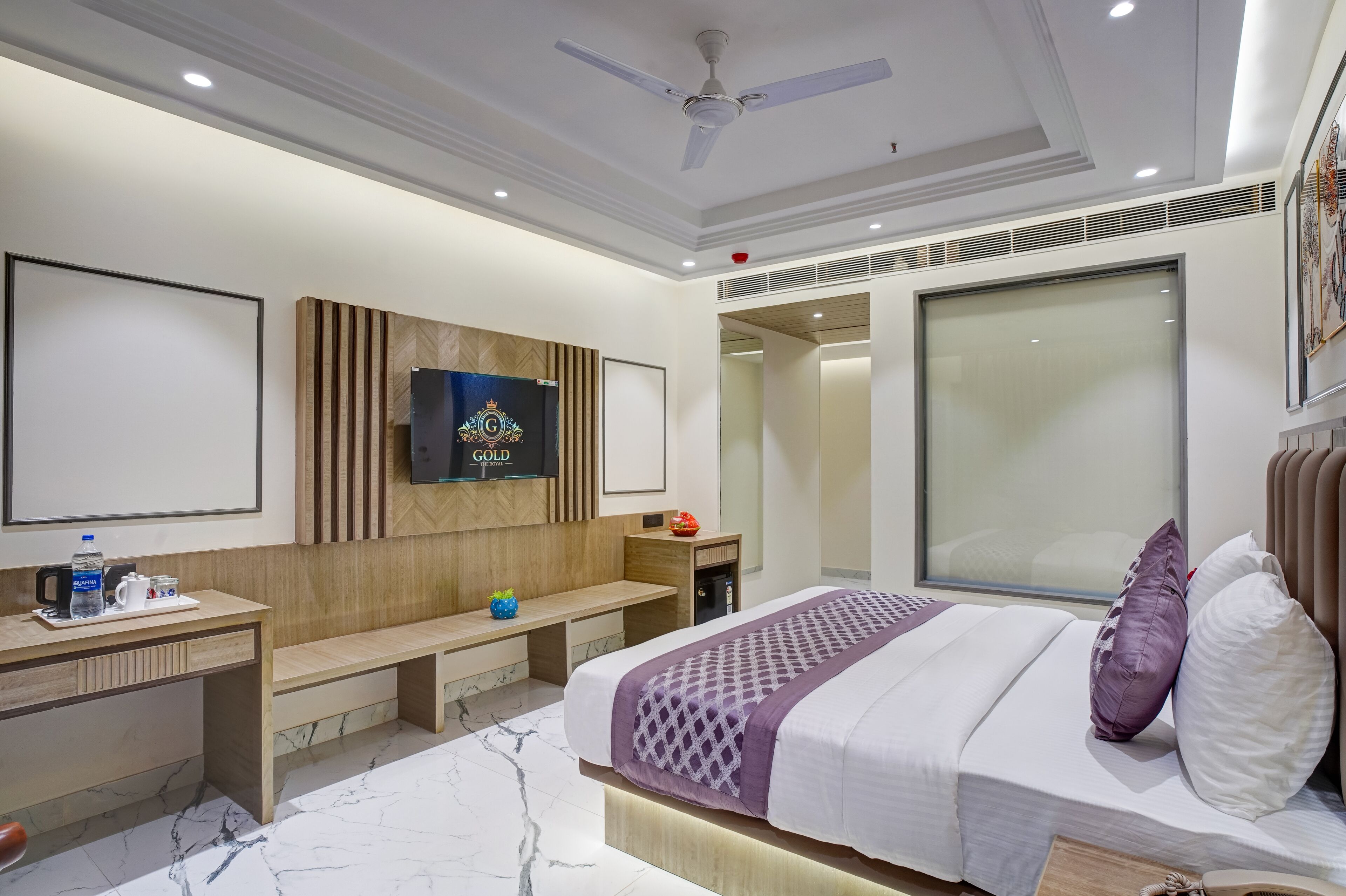 A modern hotel room with a large bed, a desk with a chair, a bench, a TV mounted on the wall, and a large window.