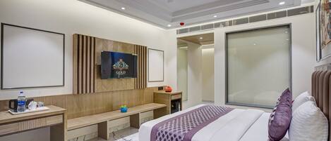 A modern hotel room with a large bed, a desk with a chair, a bench, a TV mounted on the wall, and a large window.