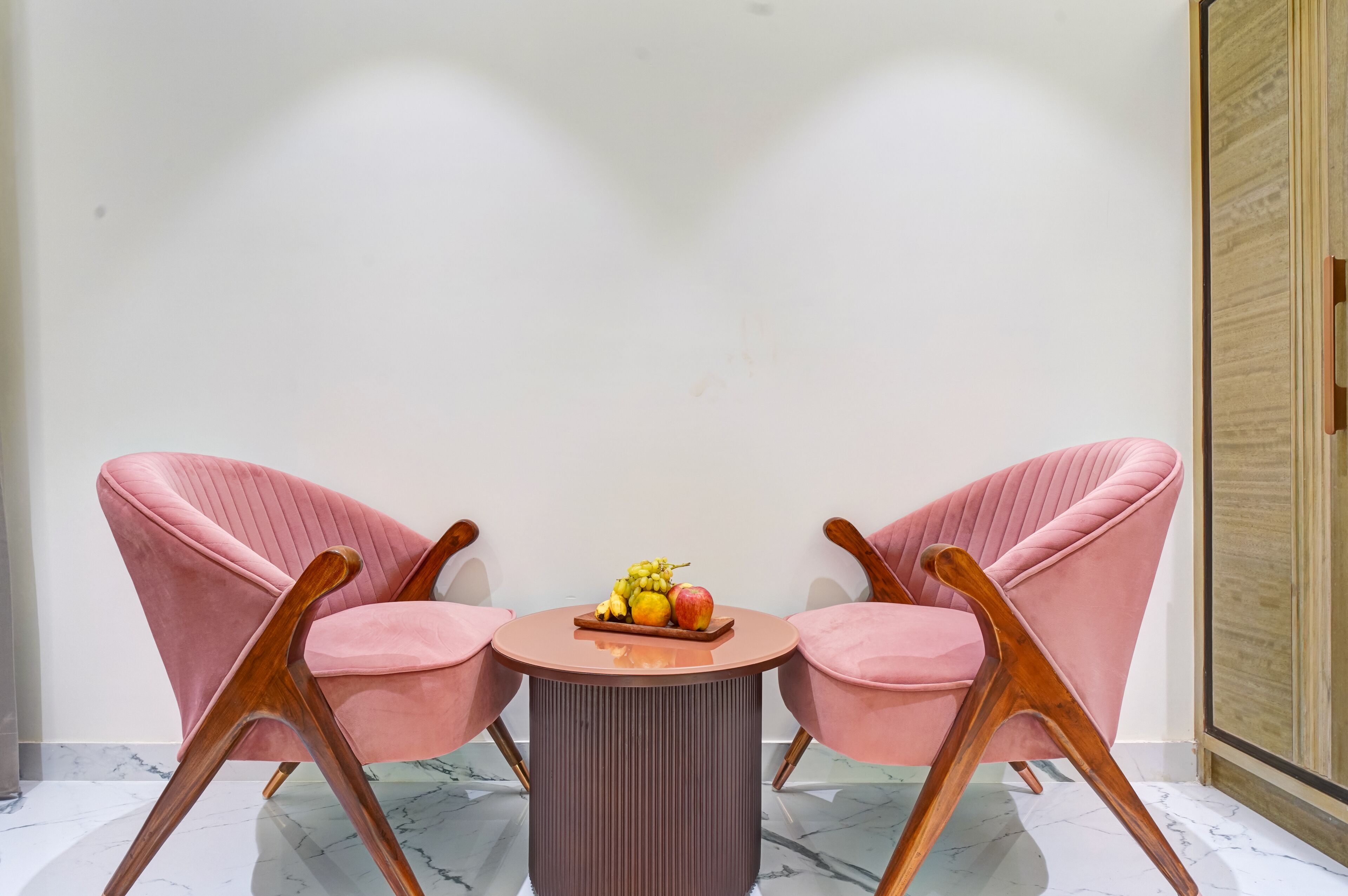 Een kamer met twee roze stoelen, een kleine ronde tafel met fruit en een houten garderobe.