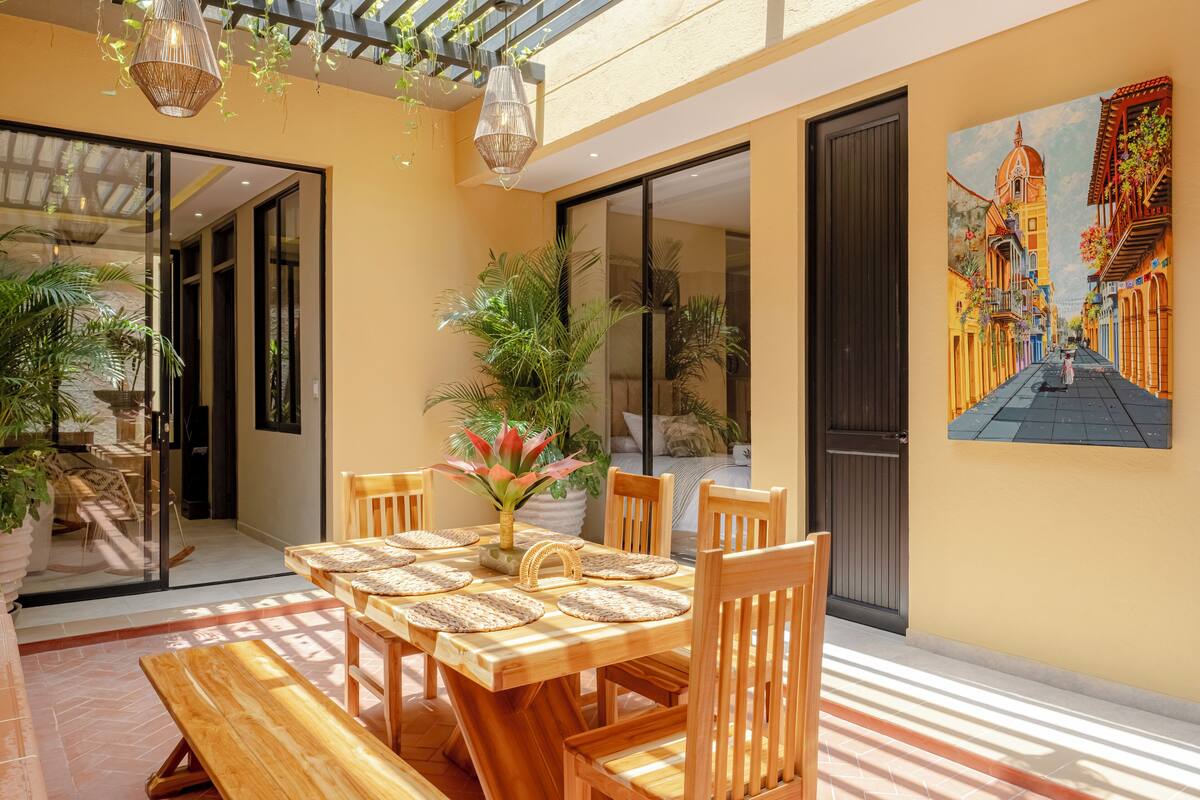 A dining area with a wooden table and chairs, surrounded by potted plants and a large window.