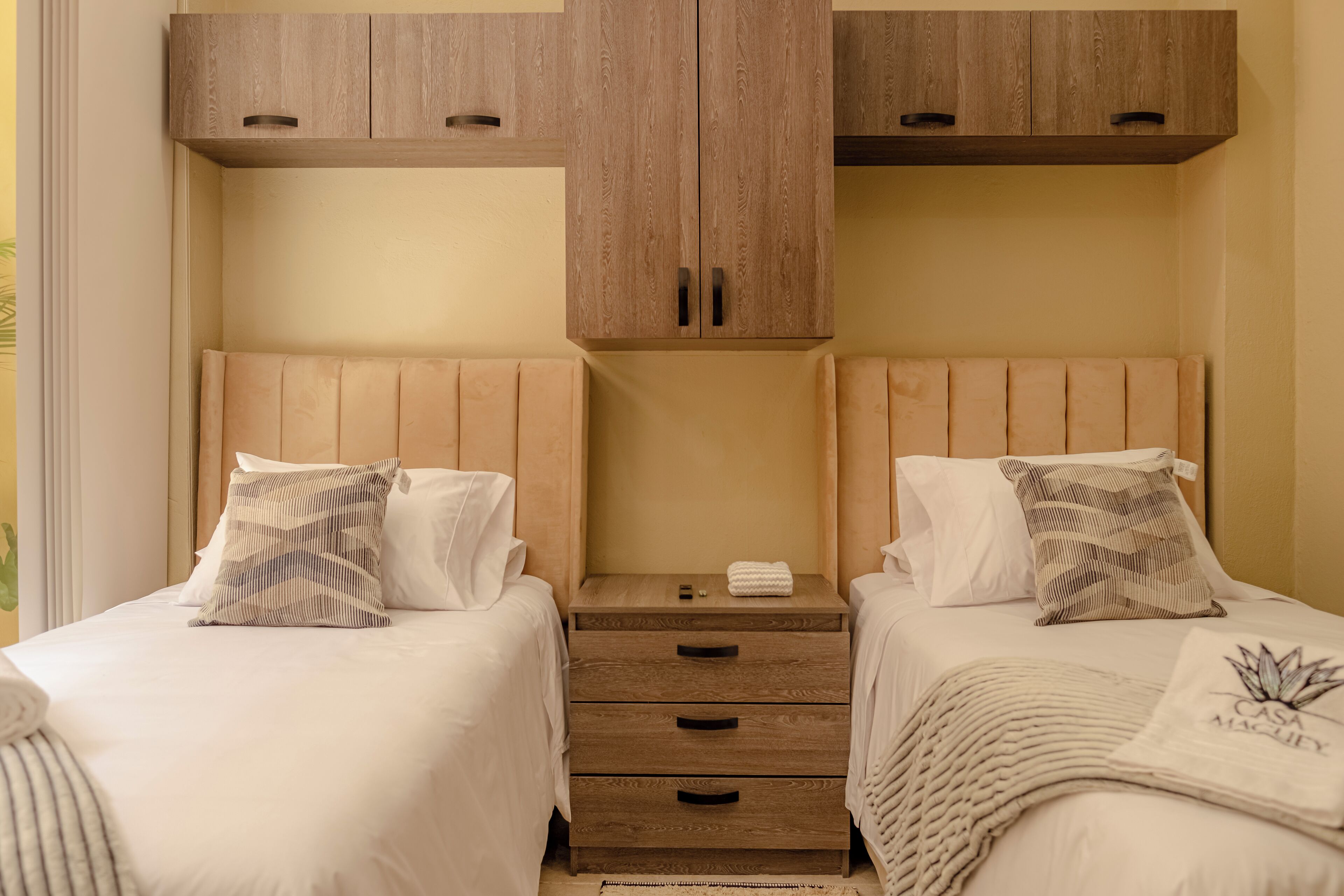 A hotel room with two beds, each with a pillow and a bedside table. There are wooden cabinets above the beds and a small wooden nightstand between them.