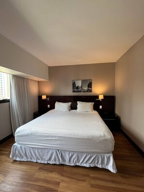 A hotel room with a large bed, two bedside tables with lamps, a window with curtains, and two wall-mounted picture frames. - Get a flat-1504 Berrini 4 estrelas (São Paulo)