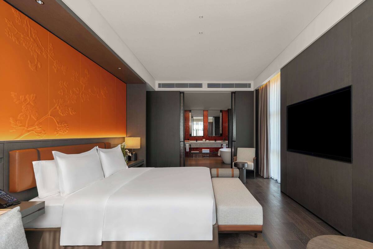 A modern hotel room with a large bed, a sofa, a TV, and a bathroom visible through an open door.