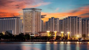 A high-rise hotel with a prominent "DOUBLETREE by Hilton" sign, reflecting in a calm body of water, surrounded by other buildings and a well-lit plaza. - Doubletree By Hilton Kaifeng (Kaifeng)