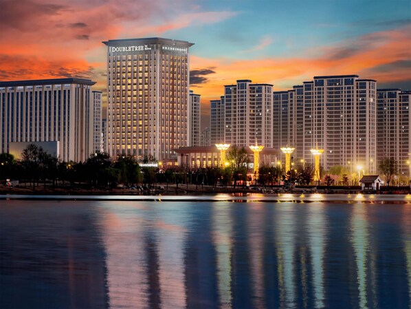 A high-rise hotel with a prominent "DOUBLETREE by Hilton" sign, reflecting in a calm body of water, surrounded by other buildings and a well-lit plaza. - Doubletree By Hilton Kaifeng (Kaifeng)