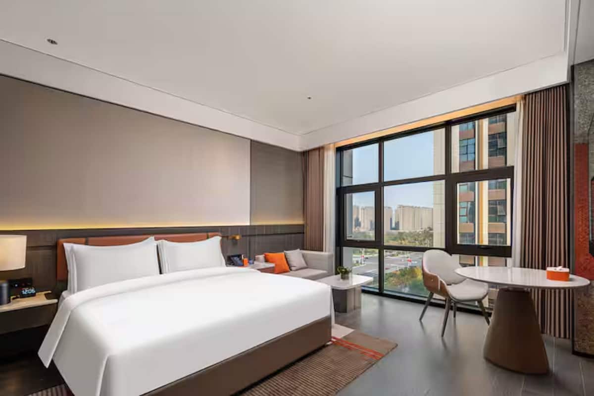 A modern hotel room with a large bed, a sofa, a small table, and a view of the city through large windows.