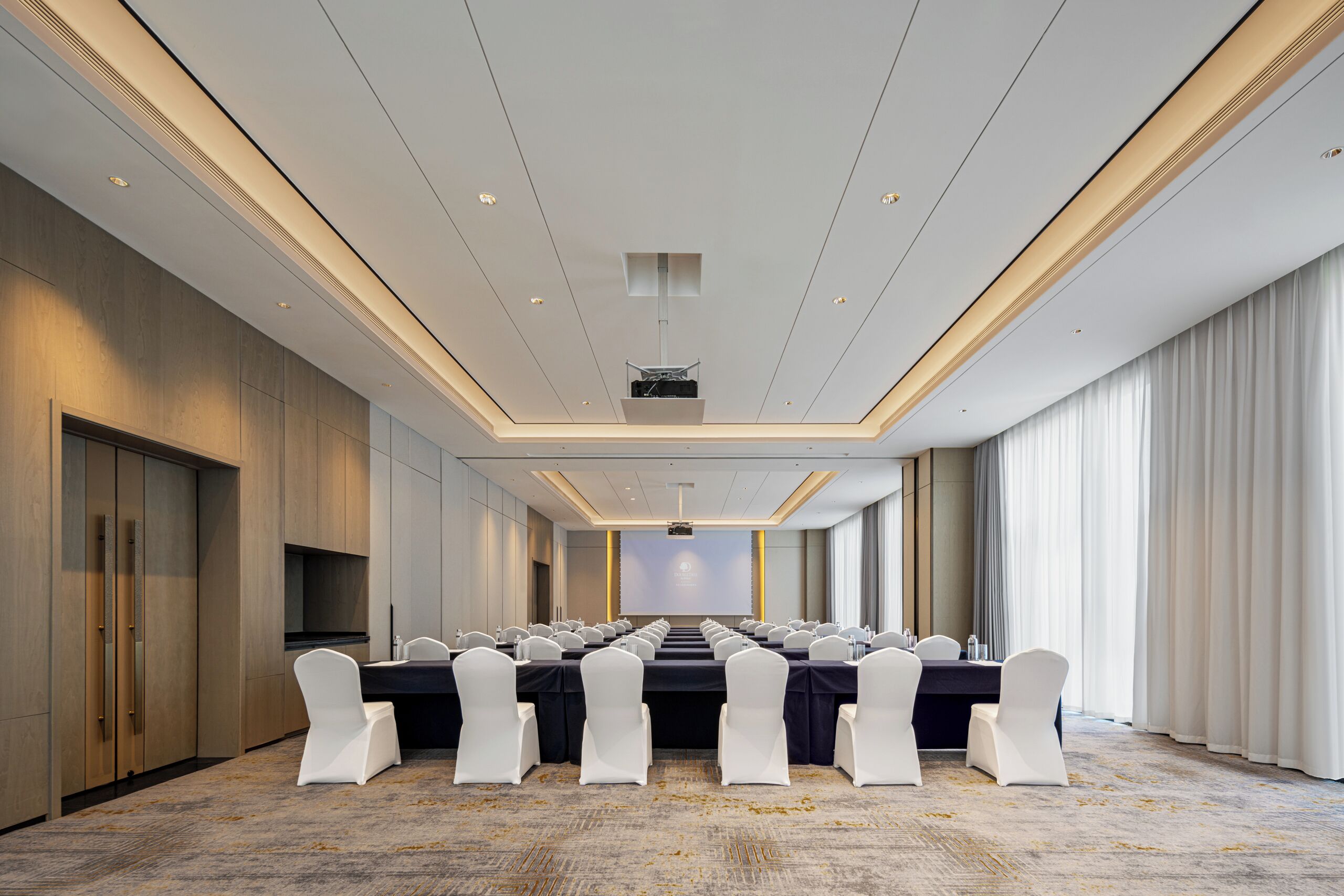 A conference room with a long table, white chairs, and a projector screen.