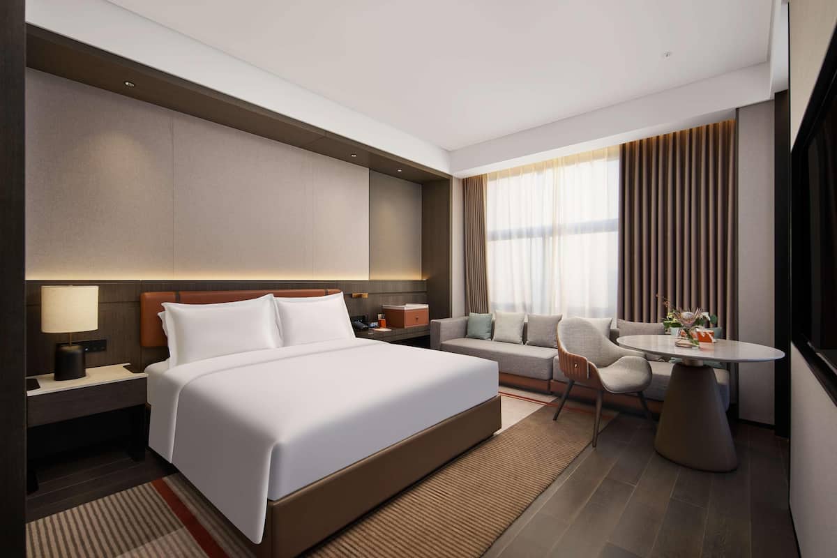 A modern hotel room with a large bed, a sofa, a dining table, and a view of the city.