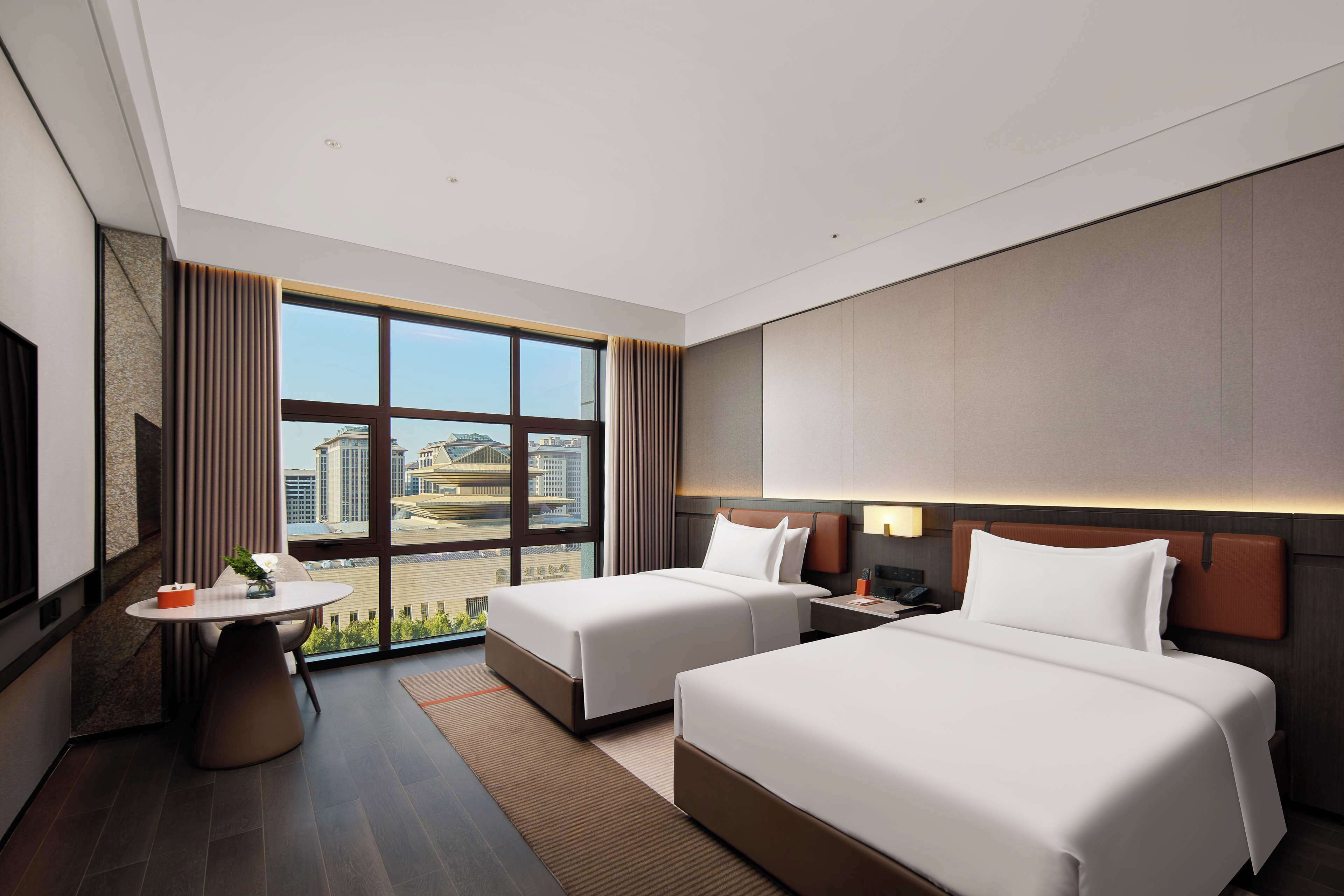 A hotel room with two beds, a desk, and a large window offering a city view.