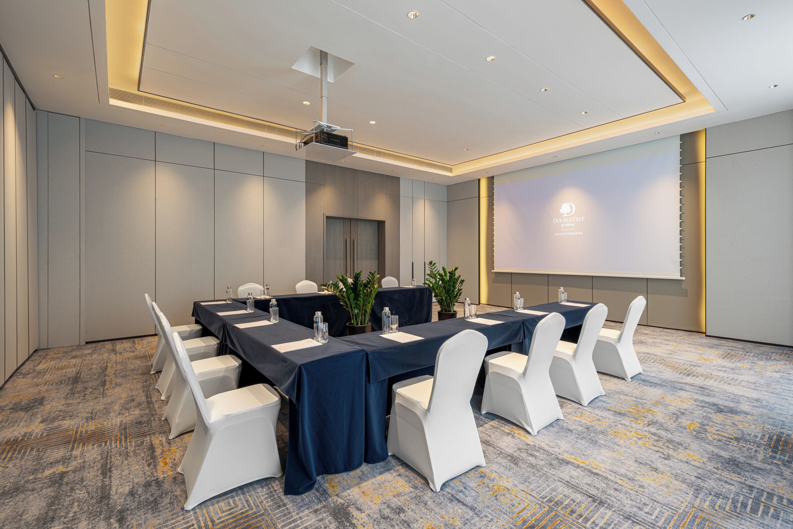 Uma sala de conferência com uma mesa comprida, cadeiras brancas e uma tela de projetor.