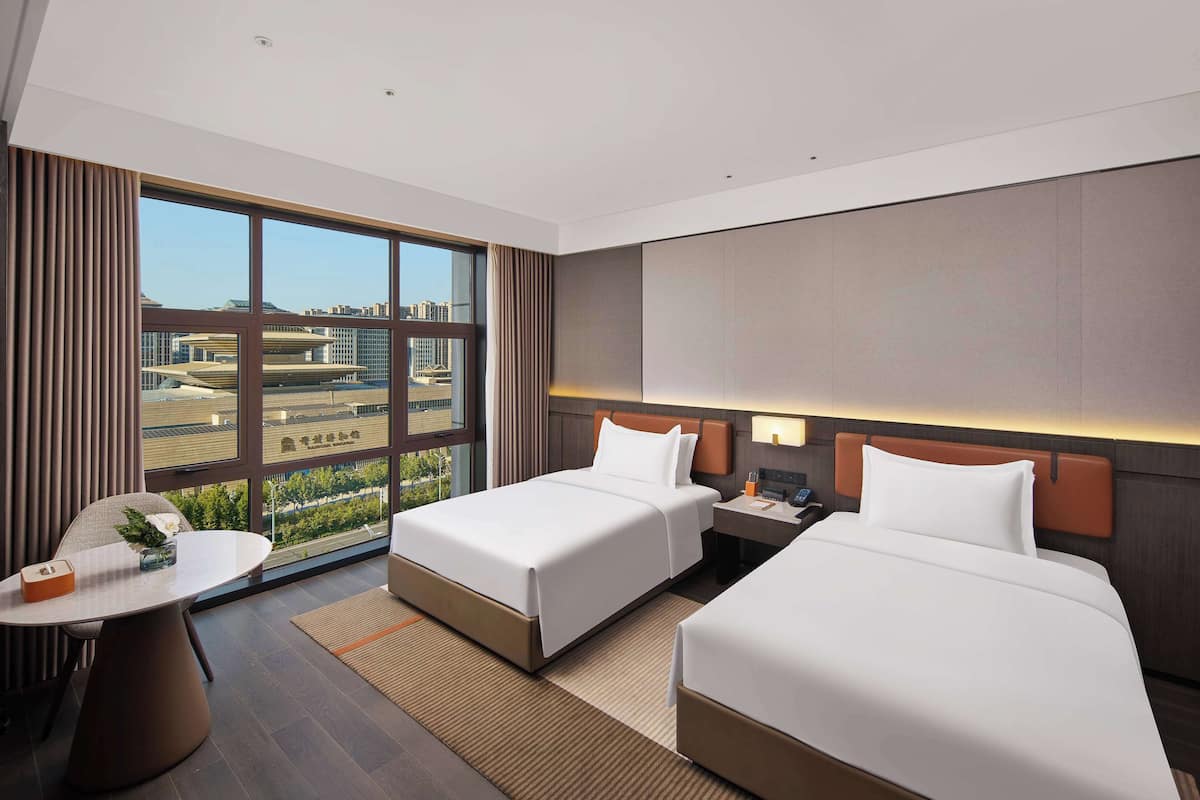 A hotel room with two beds, a desk, and a large window offering a city view.