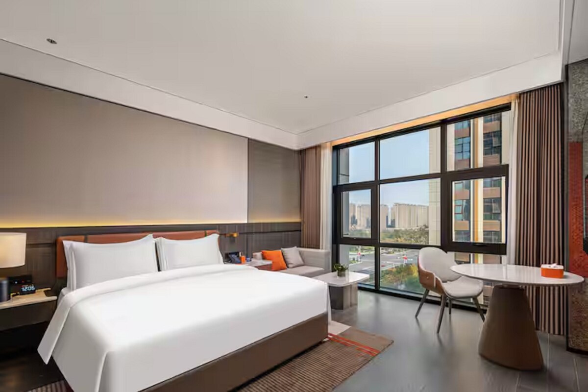 Ein modernes Hotelzimmer mit einem großen Bett, einer Sitzecke, einem kleinen Tisch und Blick auf die Stadt durch große Fenster.