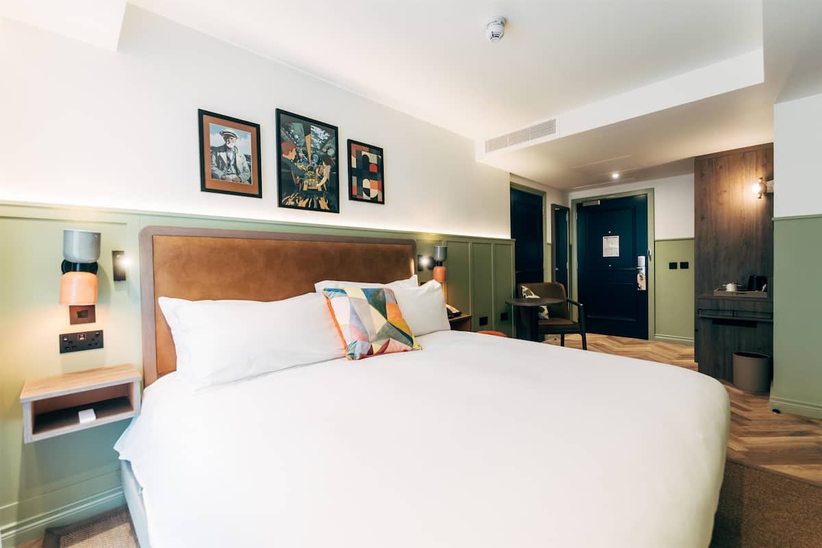 A hotel room with a large bed, a nightstand with a lamp, a wooden headboard, and two framed pictures on the wall.