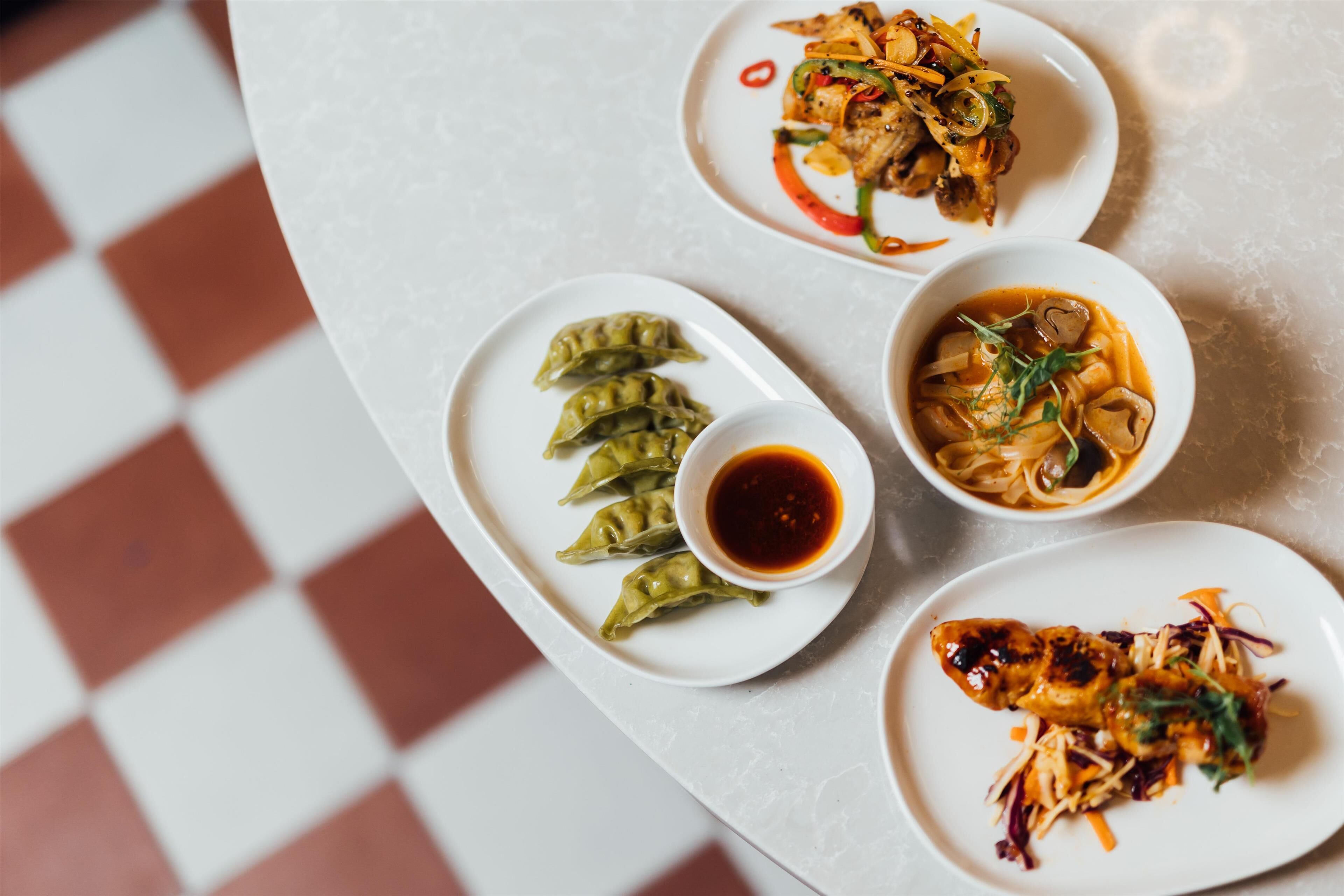 En bordopstilling med fire retter: dumplings, nudler, en kødret og en grøn suppe.