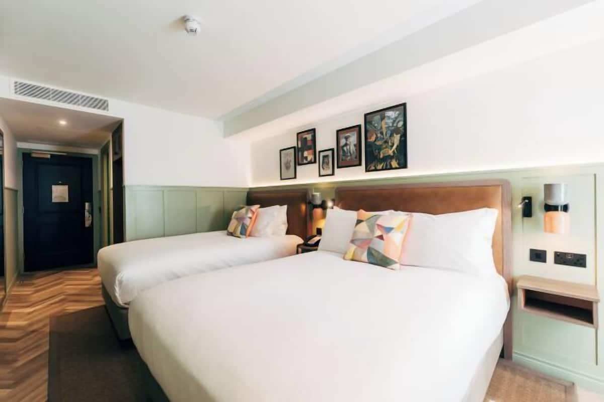 A hotel room with two beds, a headboard, wall-mounted artwork, a bedside table, and a nightstand.