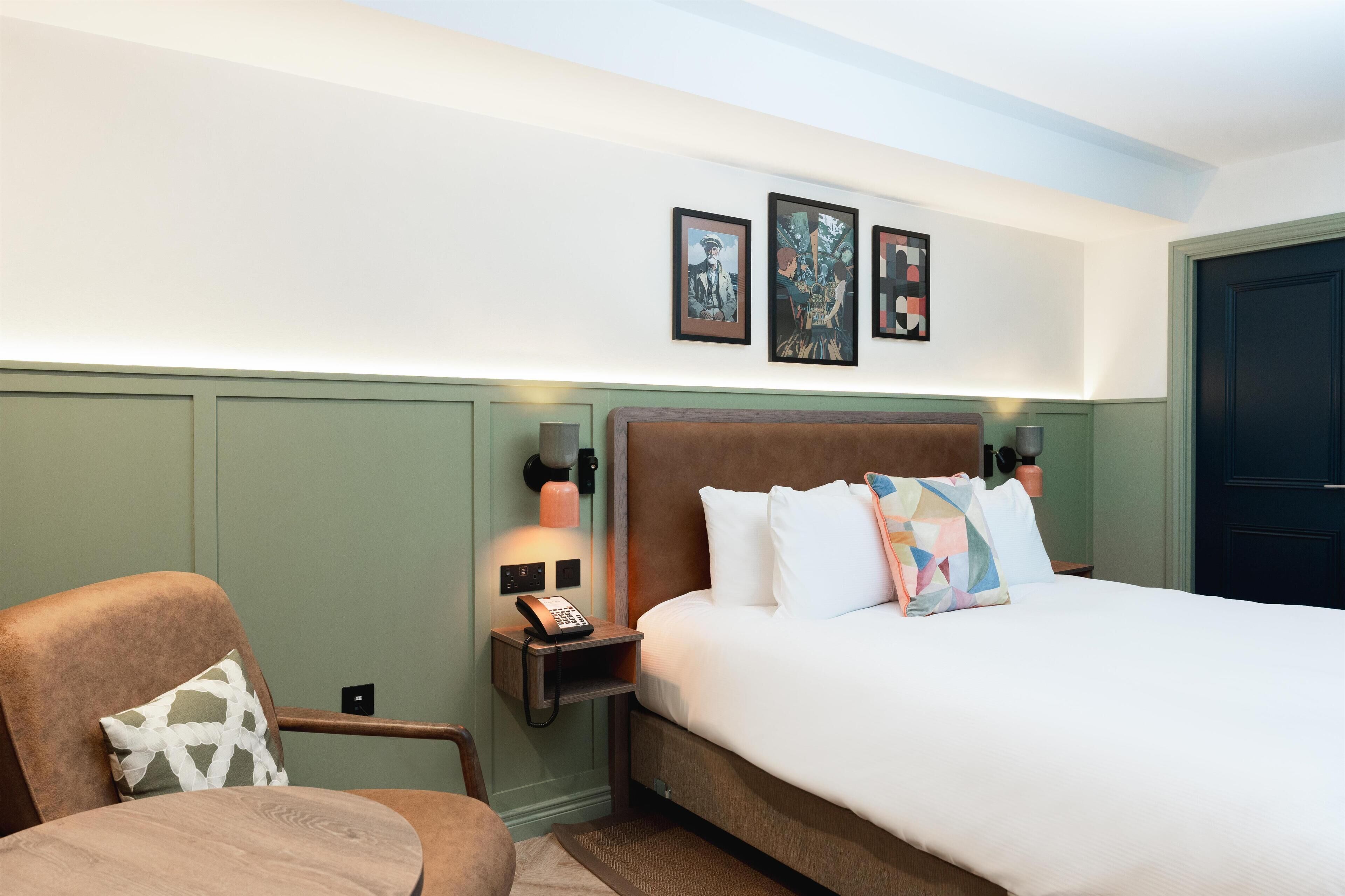 A hotel room with a bed, a chair, a nightstand with a phone, and wall-mounted artwork.