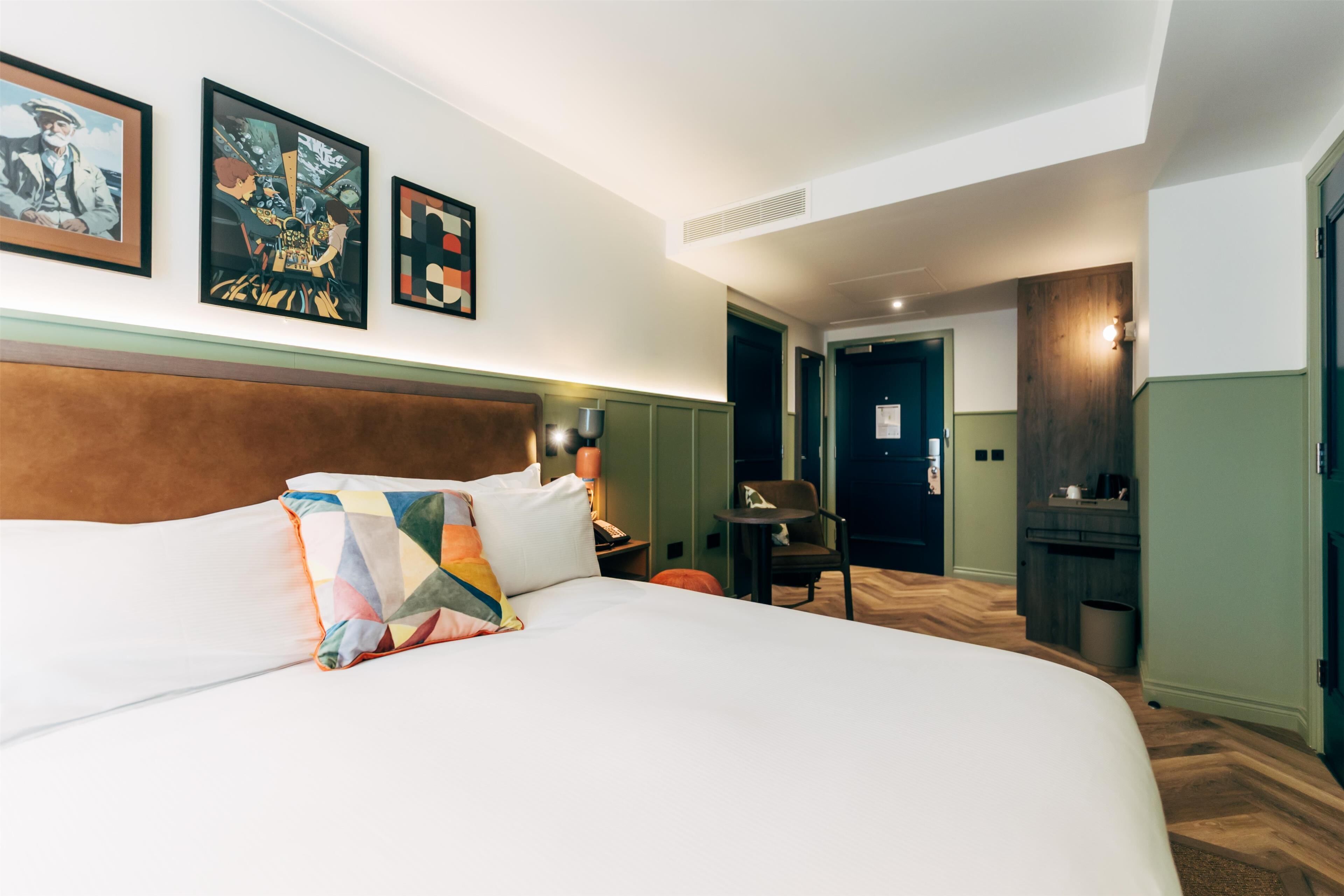 A hotel room with a large bed, a wooden headboard, a desk with a chair, and artwork on the wall.