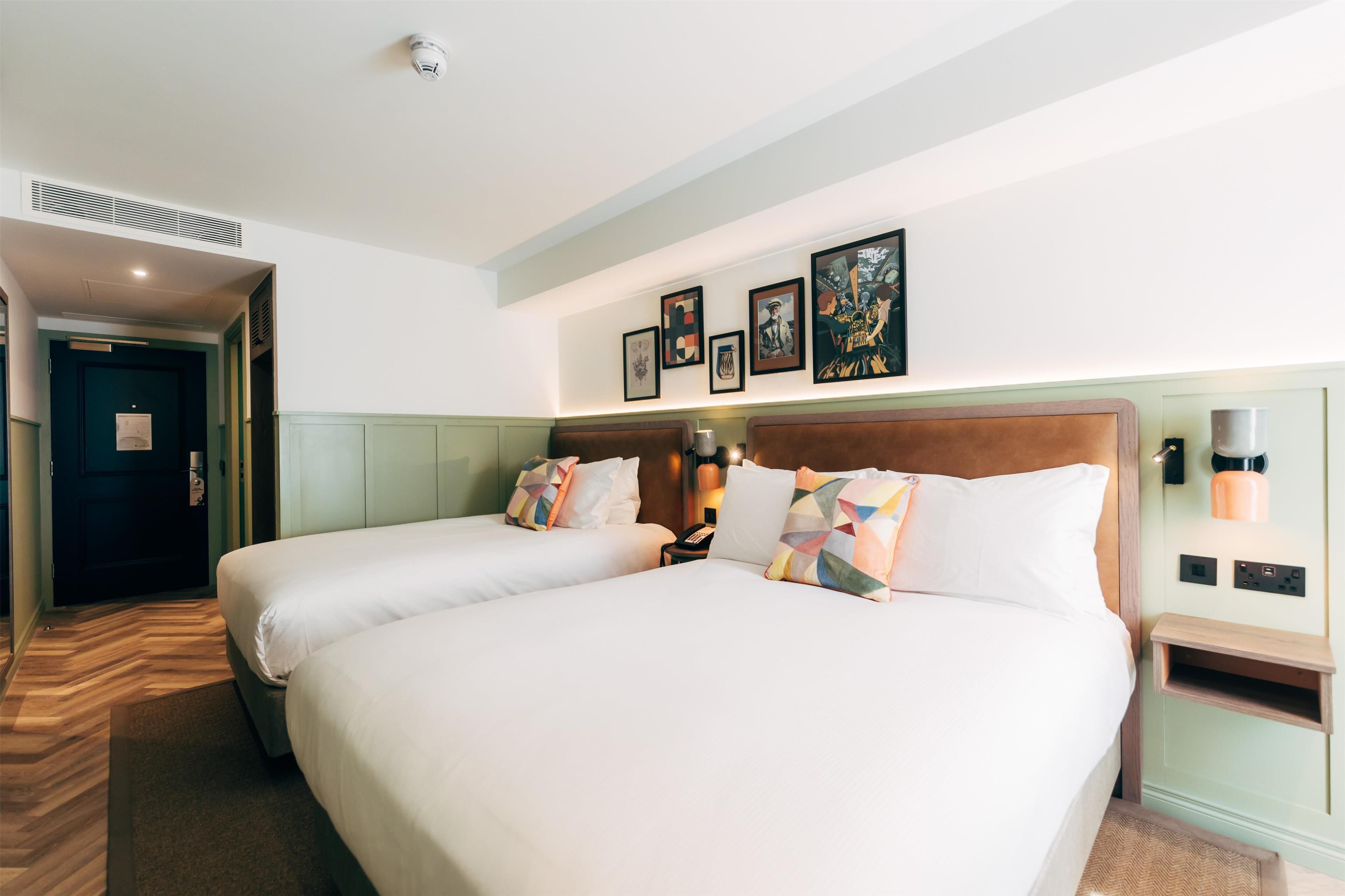 A hotel room with two beds, a nightstand, a wall-mounted lamp, and framed artwork.