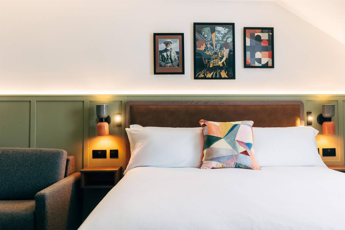 A hotel room with a large bed, two bedside tables, a chair, and three framed artworks on the wall.