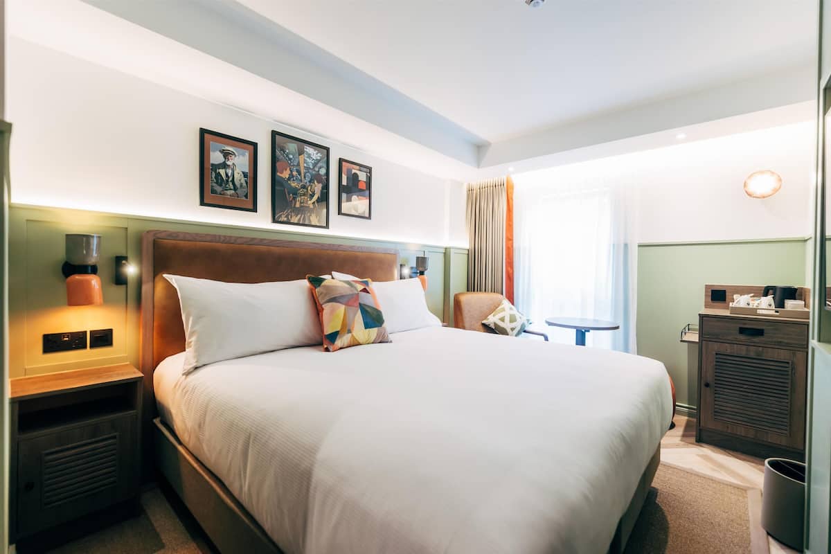 A hotel room with a large bed, a nightstand with a lamp, a small table, and a wardrobe.