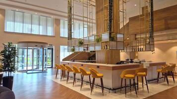 A modern hotel lobby with a central bar area, yellow bar stools, and a large potted plant.