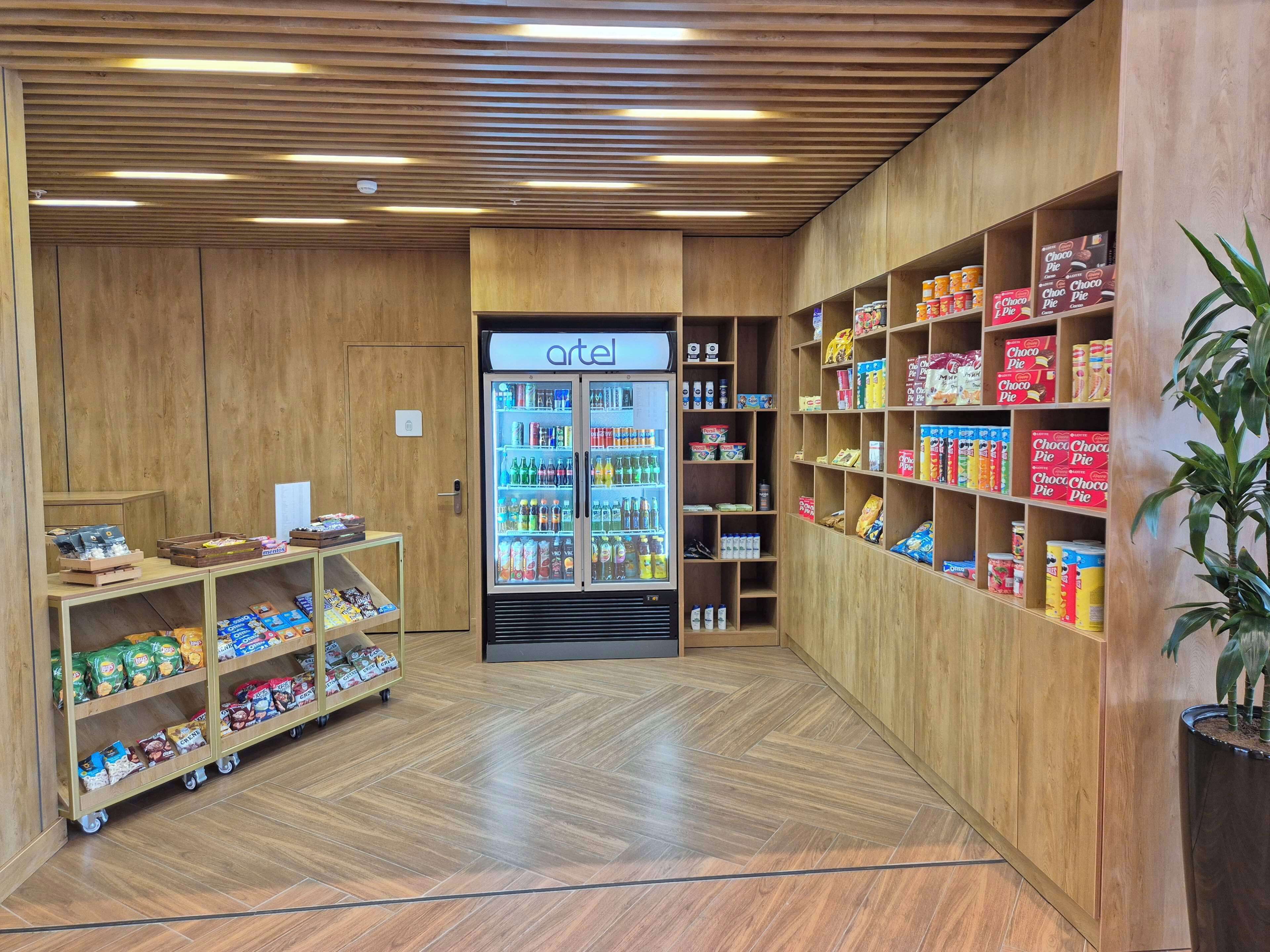 A grocery store interior with wooden walls and a refrigerated display case labeled 'artel'. Shelves are stocked with various products, and a cart with snacks is visible.