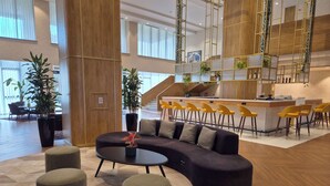 Una hall moderna con un divano curvo, poggiapiedi, un’area bar con sedie gialle e grandi piante in vaso.