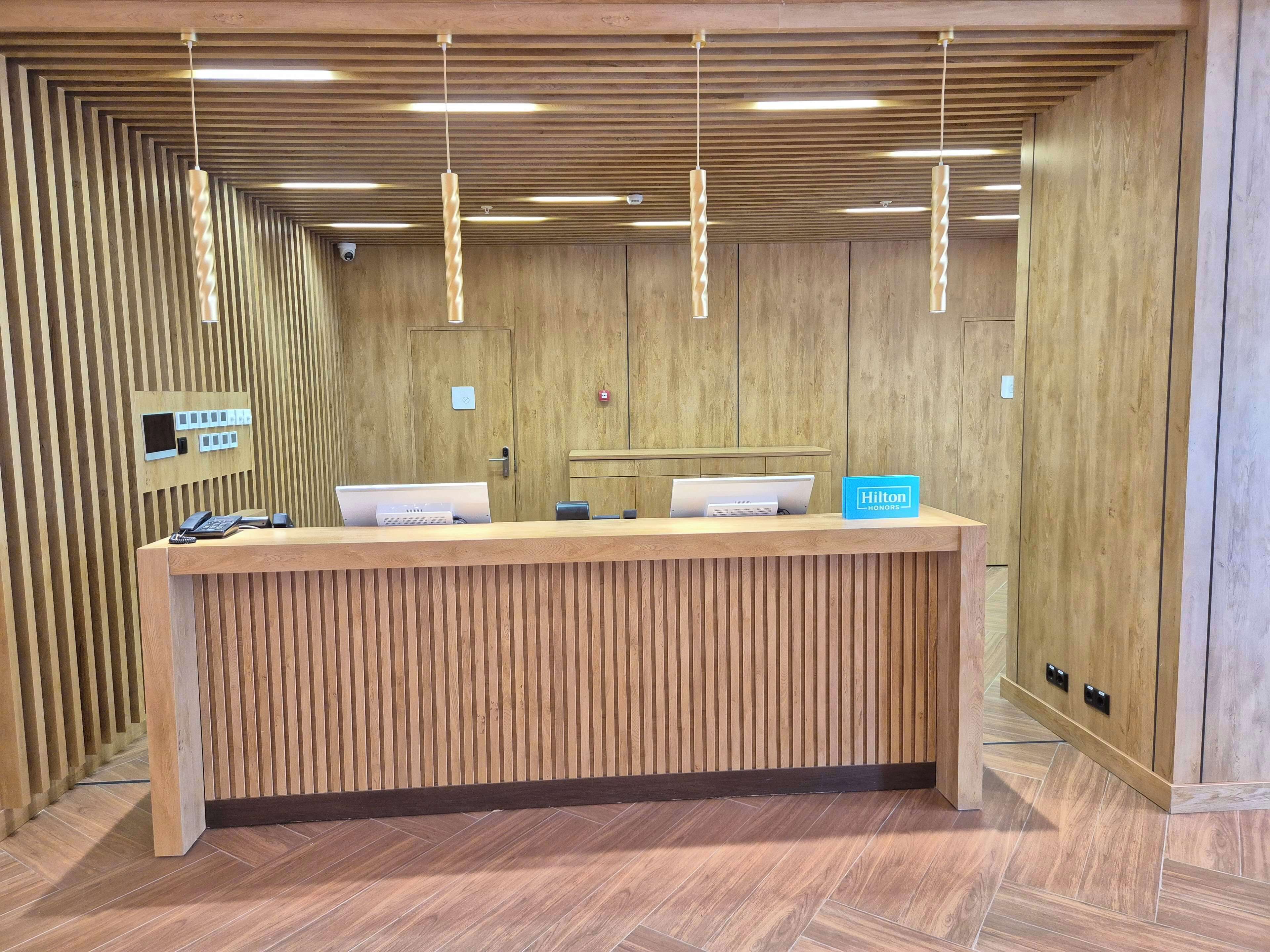 A hotel reception desk with a wooden finish, a blue sign indicating the brand, a phone, a computer, and a digital display.