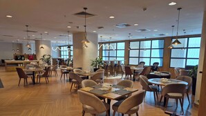 Un'ampia sala ristorante con numerosi tavoli e sedie, grandi finestre e luci a soffitto.